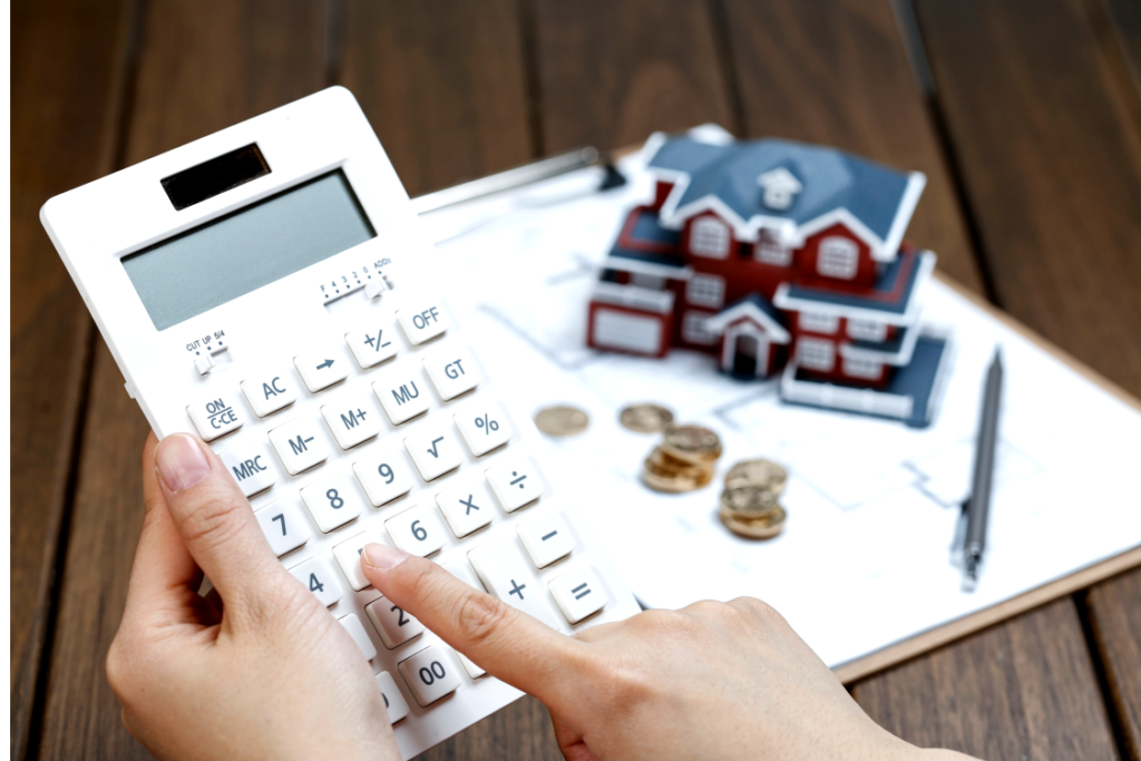 Mão segurando calculadora branca. Ao fundo, sobre uma mesa, uma casa em miniatura com moedas por perto